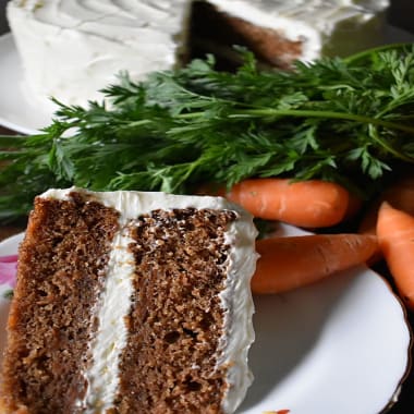 Slice of Classic Carrot Cake on a plate surrounded by carrots with green leaves